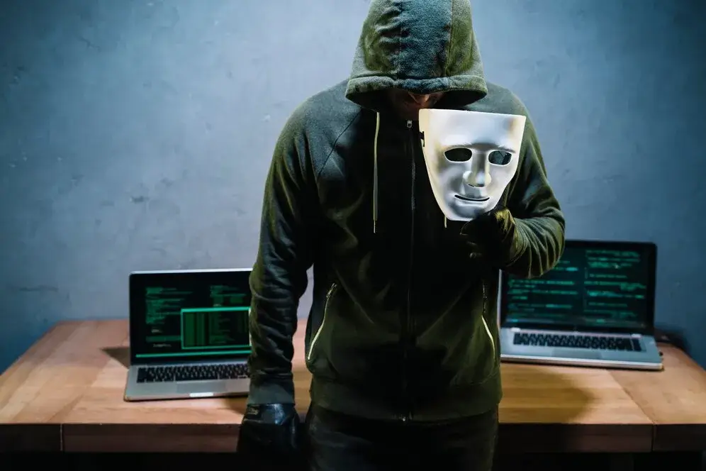 Crypto cyber threats are rising—an image of a masked hacker in front of laptops symbolizes the risk of stolen digital investments.